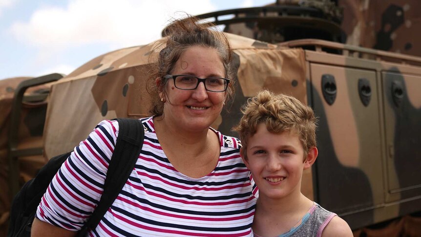 A woman and her son stand arm in arm in front of an army truck.