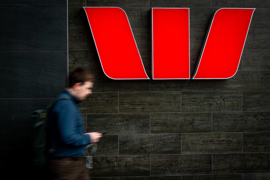 A red illuminated Westpac bank logo on a black wall with a blurred man walking past