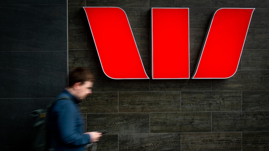 A red illuminated Westpac bank logo on a black wall with a blurred man walking past