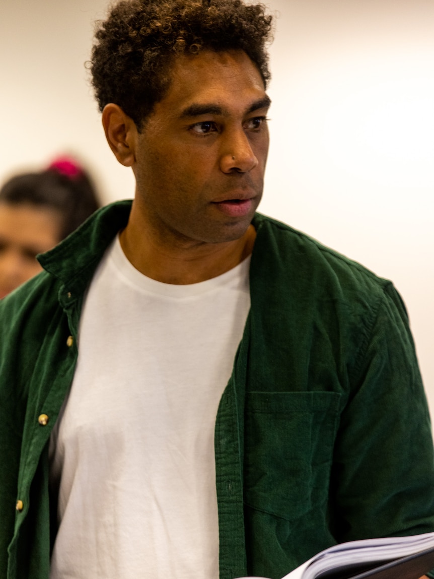 Young indigenous man with short curly dark hair wearing white tee shirt and green button-down, holding script.