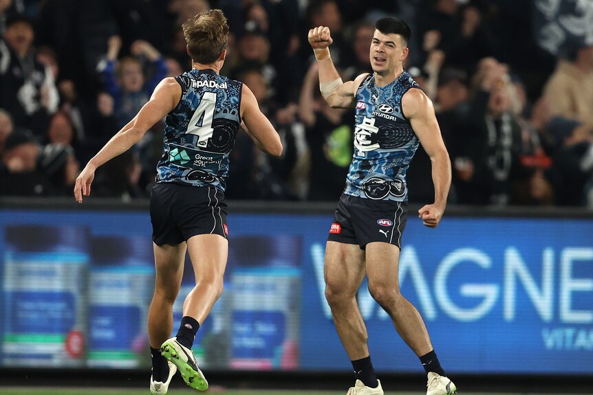 A Carlton AFL player smiles and clenches his fist in celebration as a teammate comes over to congratulate him on his goal.