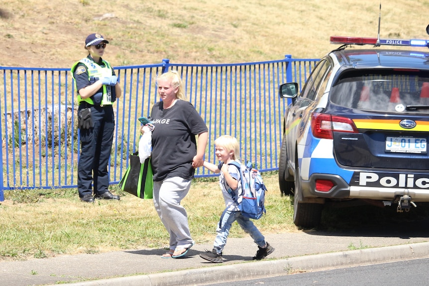 A mother and her child walk along a footpath with a police officer and police car in the background.