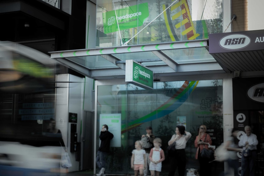 A green sign for a mental health clinic hangs above a bust stop, a bus approaches and young people stand waiting for bus.