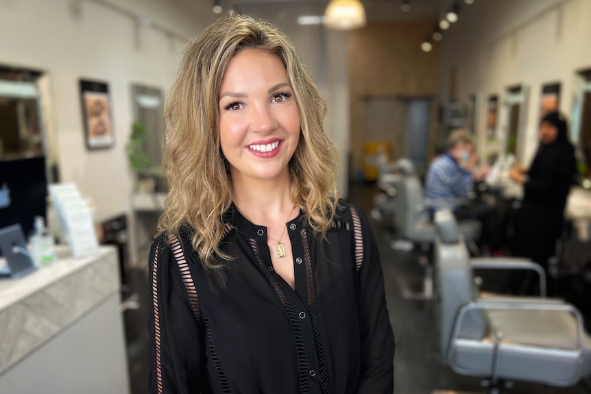 A woman with long dark blonde hair wearing a black top is in a beauty salon, smiling at the camera.