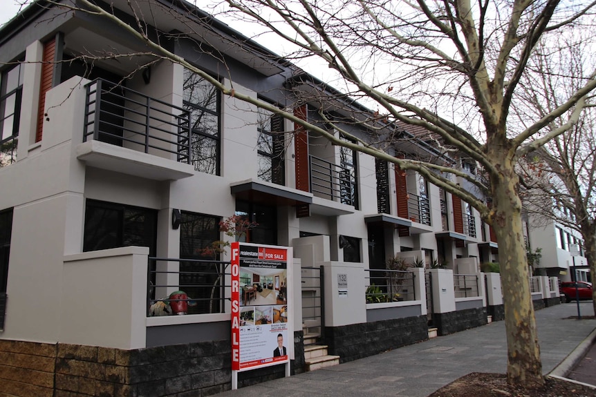 A line of townhouses, one with a for sale sign in front. The trees are bare and the sky is grey.