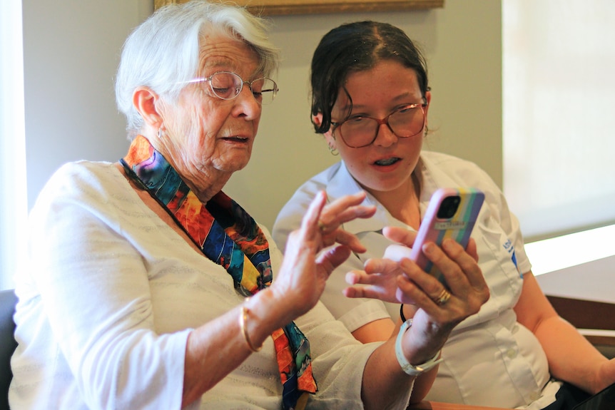 An older woman attempts to use a smartphone while a teenager girl looks on and offers advice.