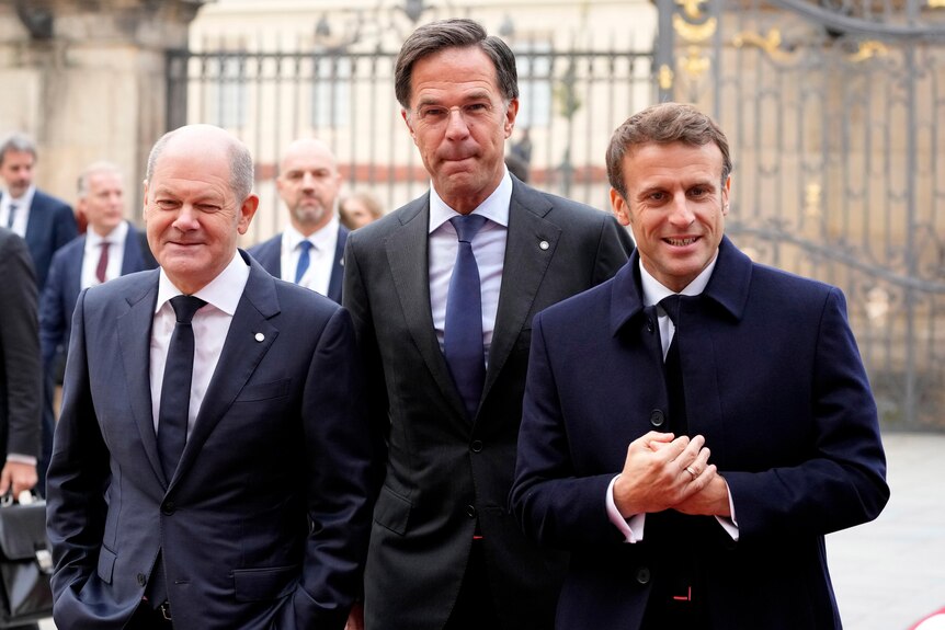 Three men in dark suits, one of them France's president, Emanuel Macron, walk towards the camera 