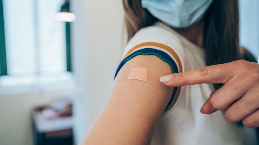 Woman wearing face mask and pointing at her arm with a bandage after receiving COVID-19 vaccine.