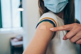 Woman wearing face mask and pointing at her arm with a bandage after receiving COVID-19 vaccine.