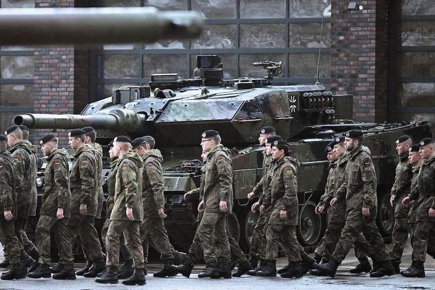 Tank crew members in battle fatigues walk past a parked tank.