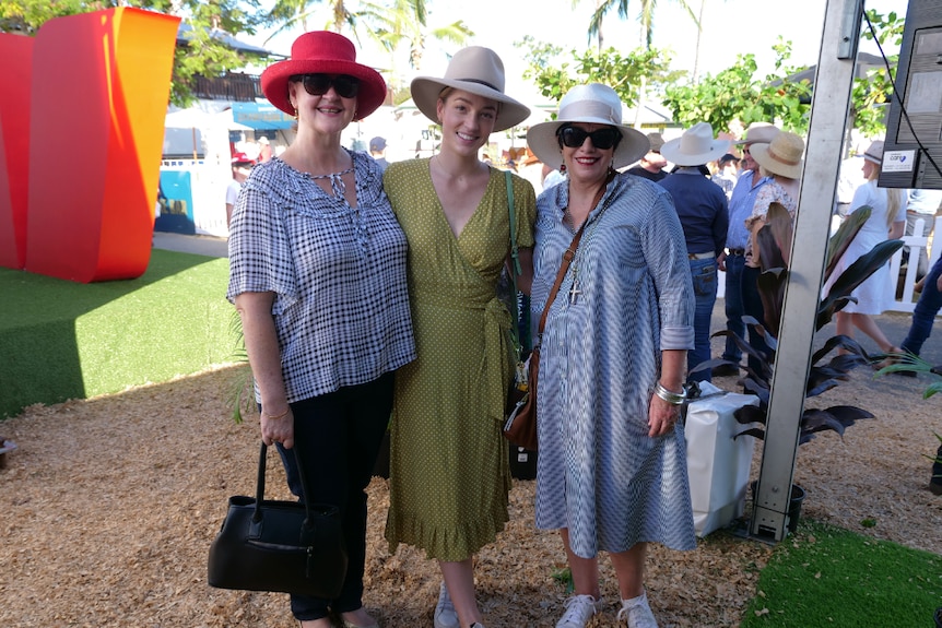 Three women stand smiling at the camera, people in background.