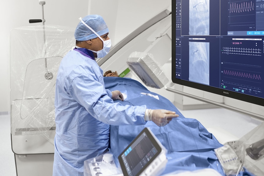 Man in blue surgical scrubs, mask, cap, has person lying on an imaging equipment, watches large screen, small monitor on left.