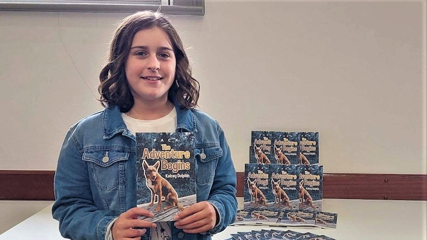 A young girl smiles while holding a book. Other copies of it are stacked on a table behind her.