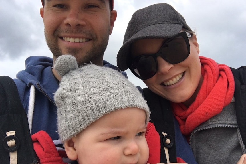 Sam, Renee and baby Hamish smile at the camera in a family photo outside.