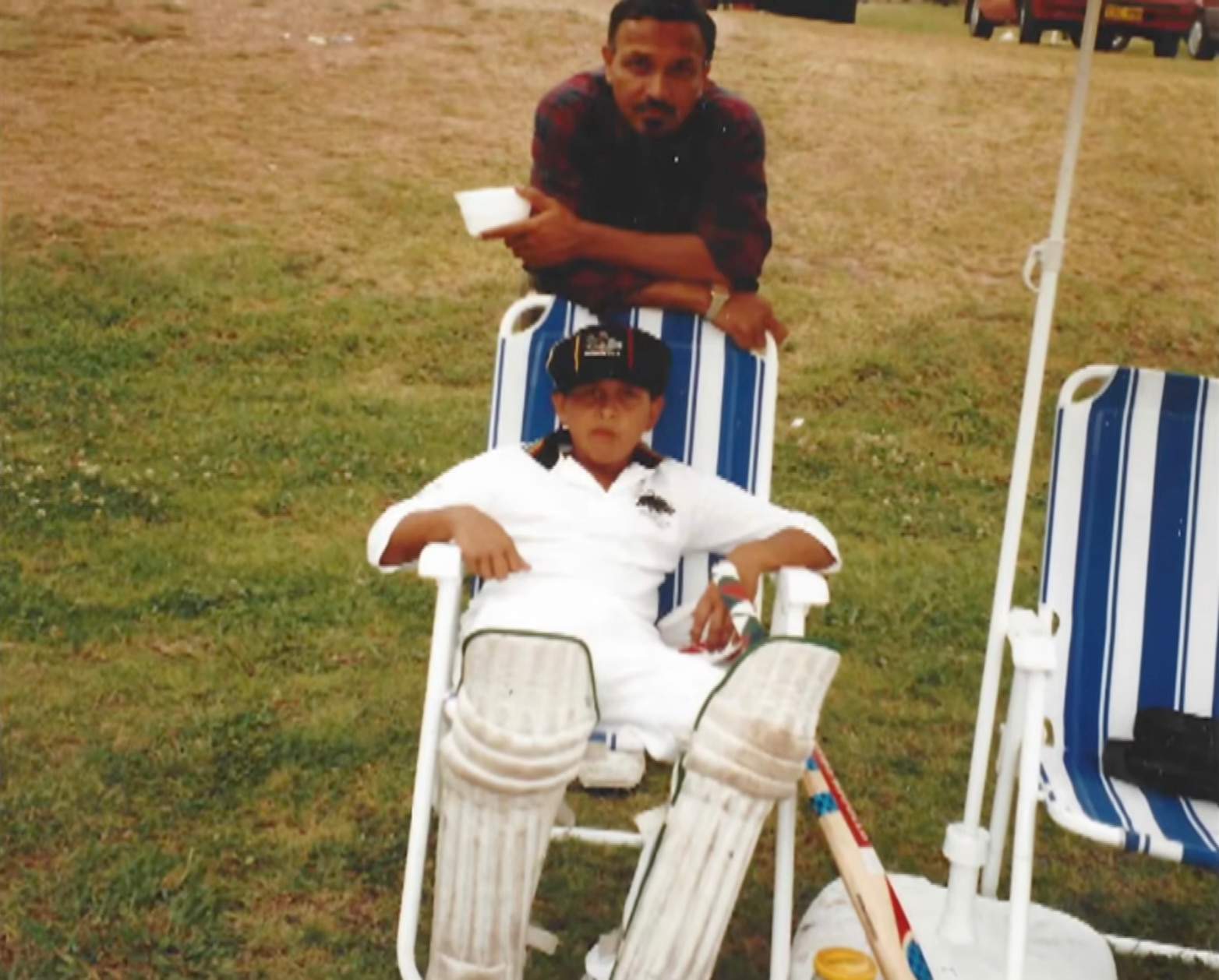 Usman and Tariq Khawaja on the grass. Usman is seated and wears cricket whites while Tariq leans over the top.
