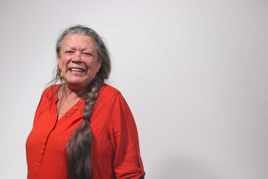 A woman wearing a red shirt standing against a blank wall.