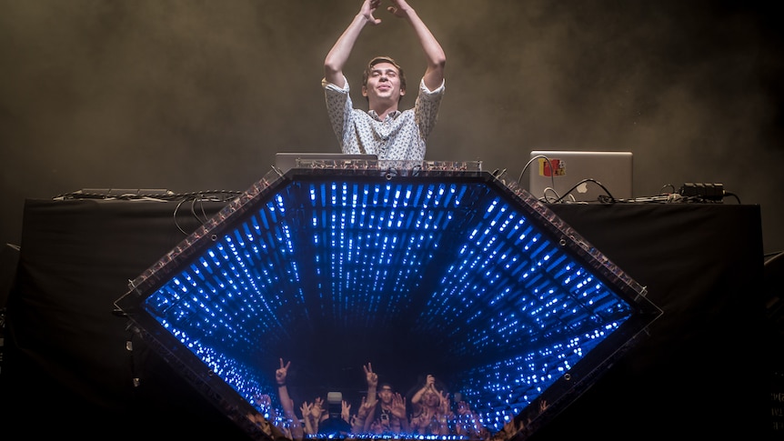 Flume raises his hands, clapping behind his hexagonal stage production which reflects faces of the crowd