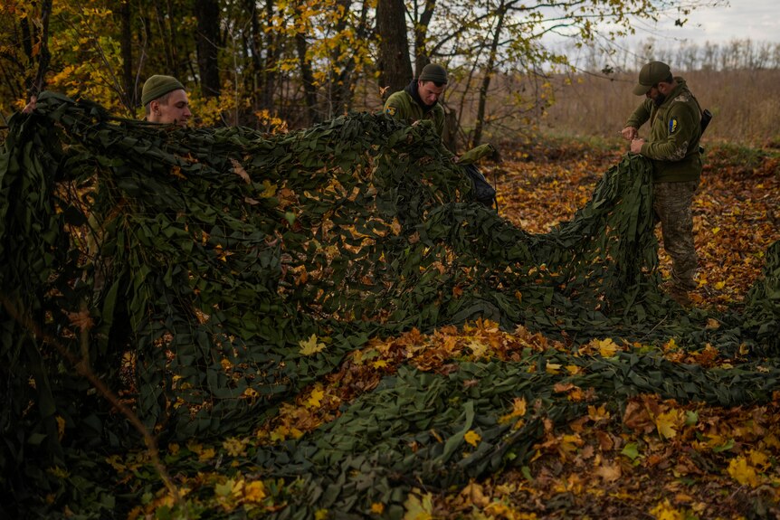 Three men dressed in camouflage hold up a large green mesh net in a forest