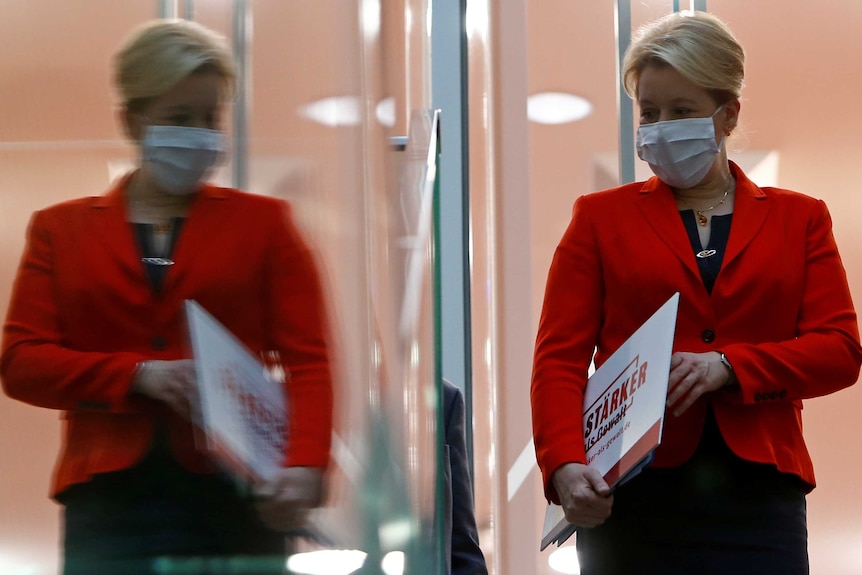 Franziska Giffey's reflection is cast on a window as she walks while wearing a face mask and carrying a sign