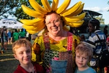 A woman wearing a banana head band stands smiling with her two kids.