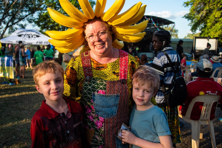A woman wearing a banana headband stands smiling with her two children.