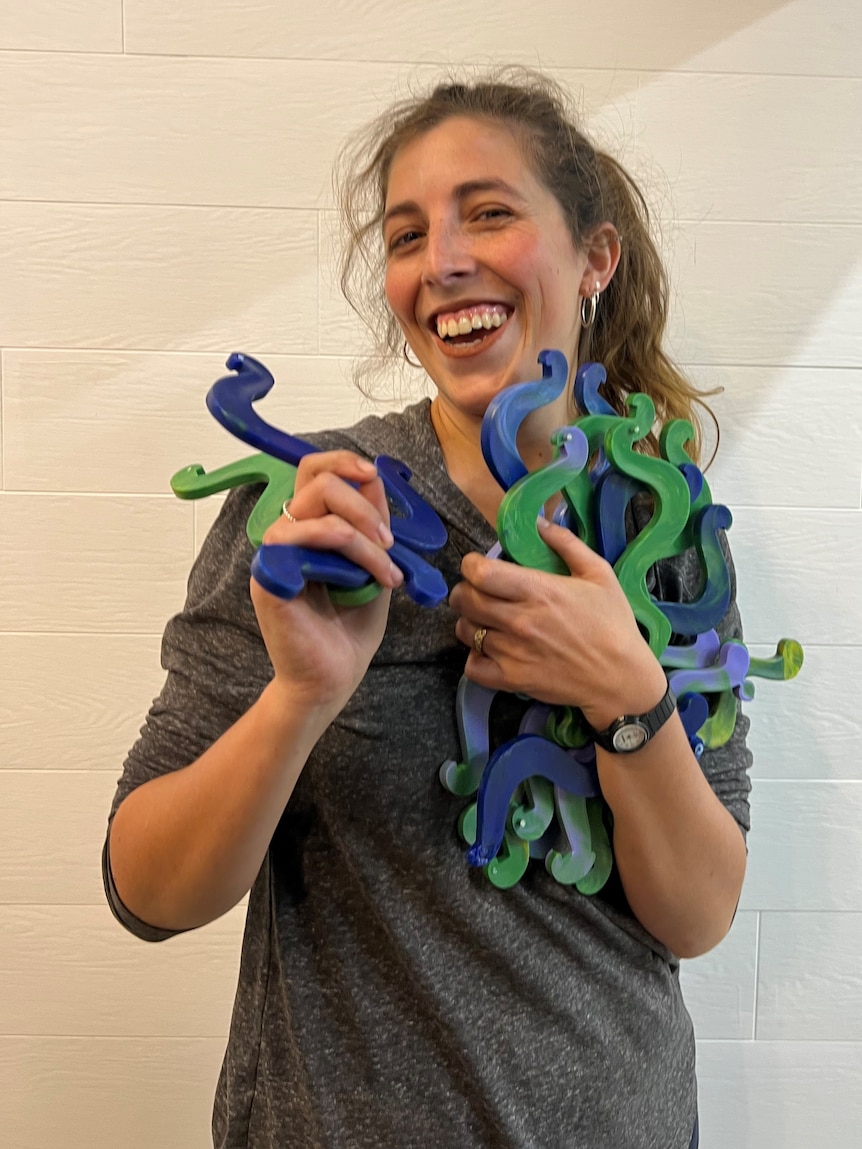 Young woman smiles broadly and holds multiple squiggly pieces of plastic -they are iPad holders made from recycled bottle lids.