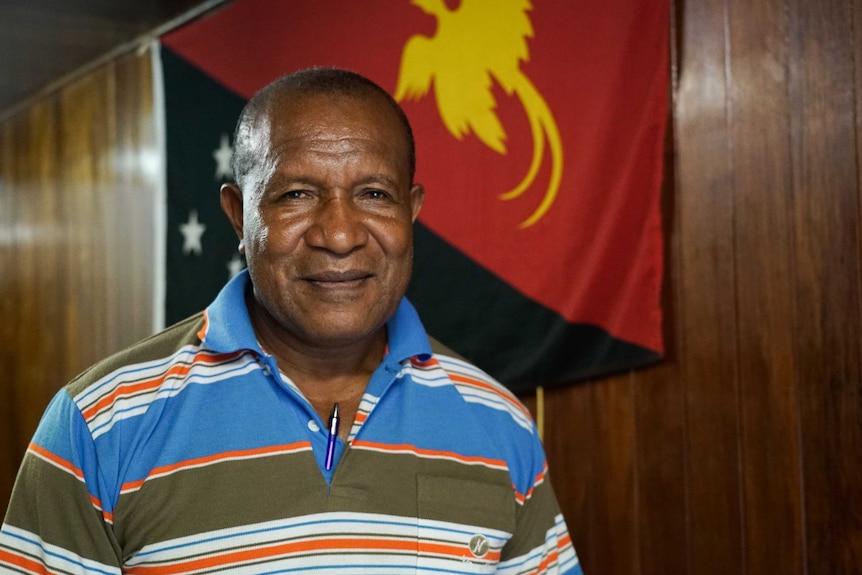 A man stands in front of a wall with the PNG flag draped on it