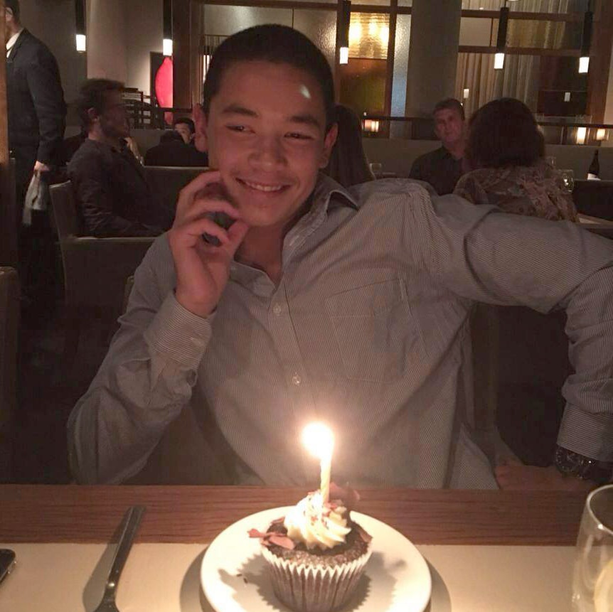 Wesley Ballantine smiles sitting at a table with a cupcake with a candle on it in front of him.