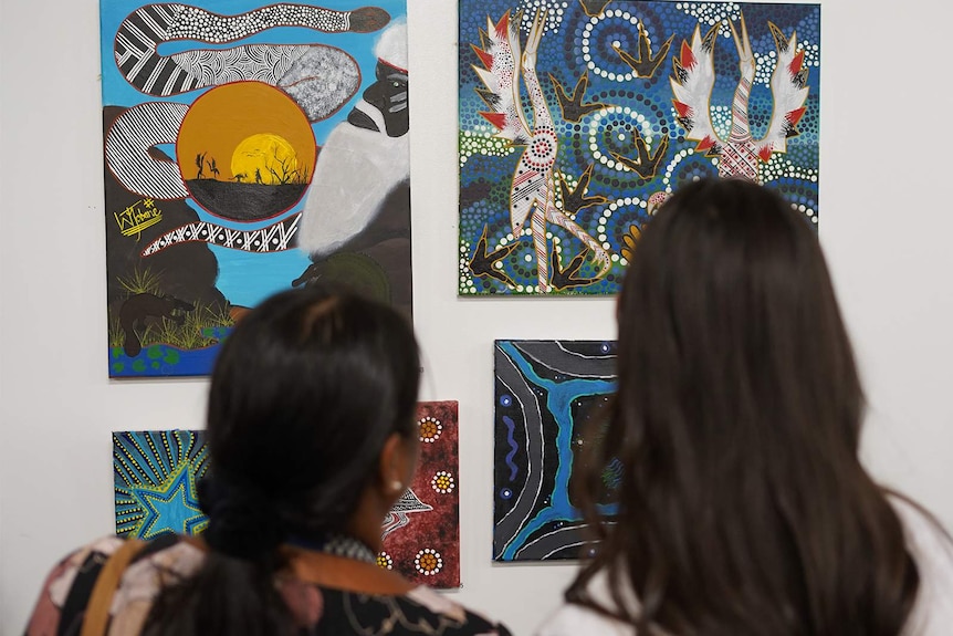 Stunning, colourful works of Aboriginal art displayed on a wall, two women looking at the art.