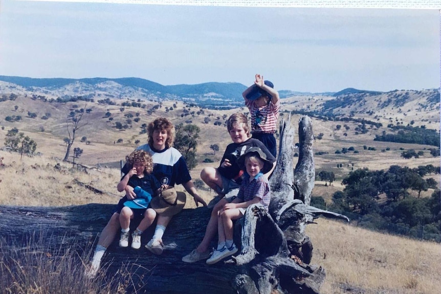 An older, faded photo of Fiona, her mum, and siblings sitting on a large tree stump in a rural area.