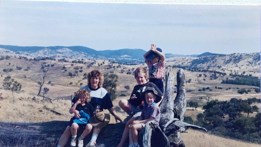An older, faded photo of Fiona, her mum, and siblings sitting on a large tree stump in a rural area.