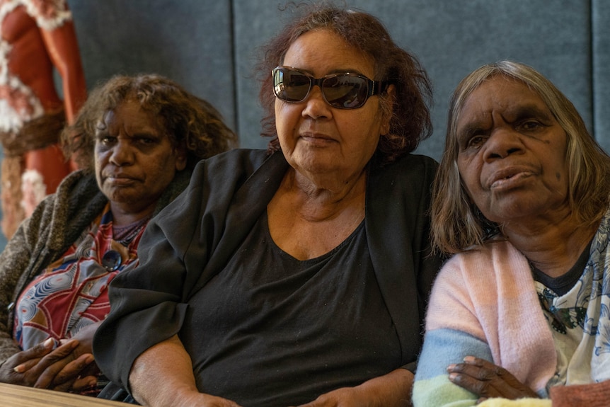 Three Indigenous women sit side by side. The woman in the middle wears sunglasses.