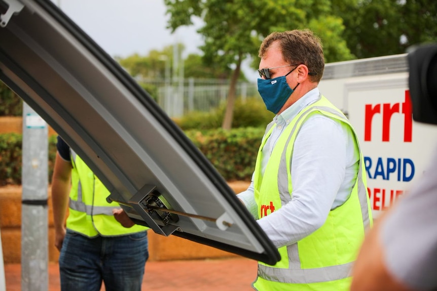 A photo of a man wearing a mask and high-vis vest closes a van boot.
