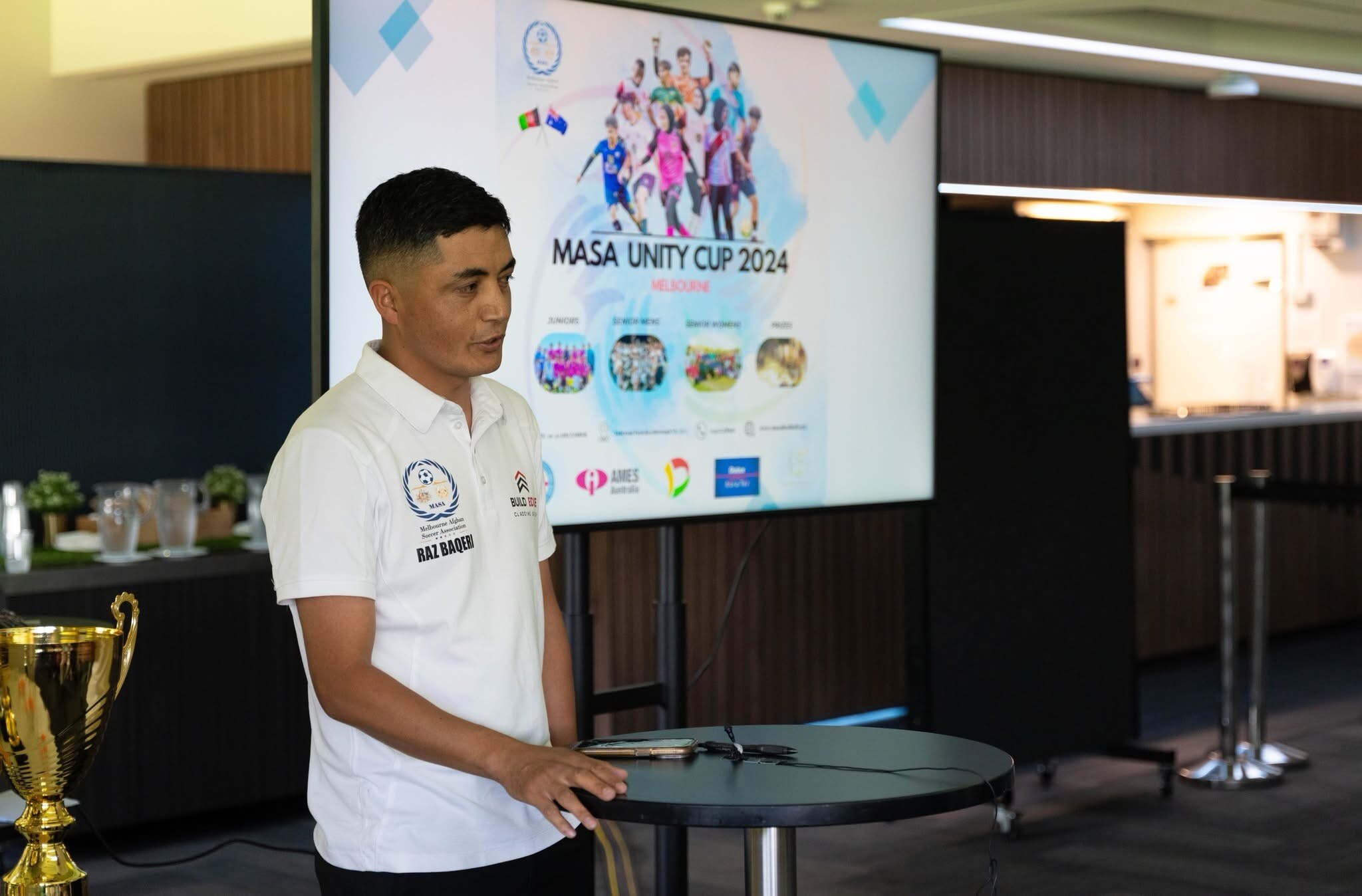 A man standing behind a table talking to people off camera with a display screen behind him