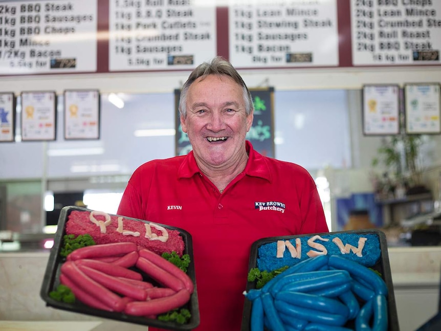 Man with beaming smile, holds two trays of sausages out, one maroon, one blue, with QLD, NSW spelt out at the top in pork rind