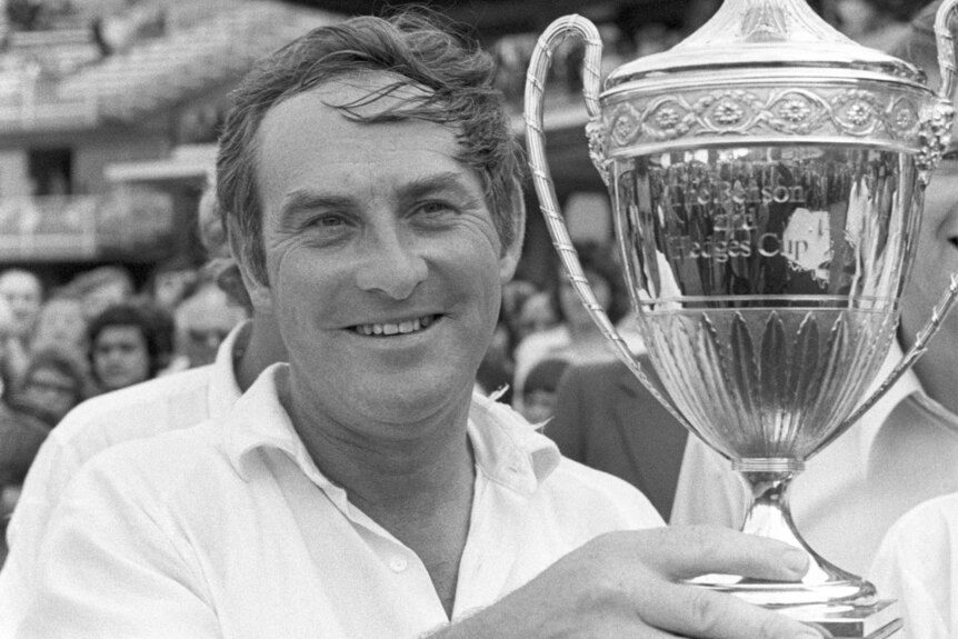 A black and white photo of cricketer Ray Illingworth in a white button-up shirt, holding up a cup-style trophy.