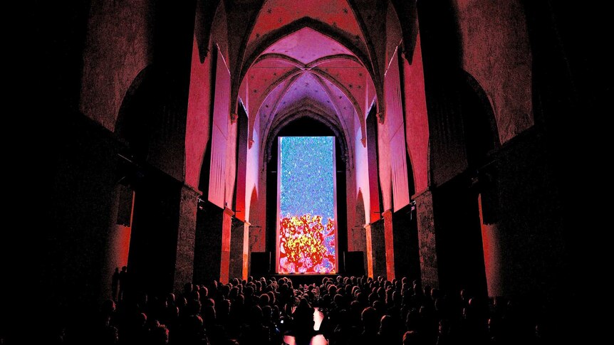 An audience watches a vertical cinema screen showing an abstract image