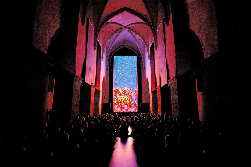An audience watches a vertical cinema screen showing an abstract image