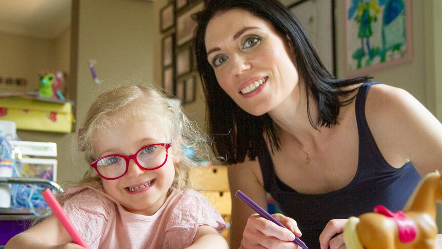 Estelle wears bright red glasses and sits at a table doing colouring in beside her mum.