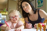 Estelle wears bright red glasses and sits at a table doing colouring in beside her mum.