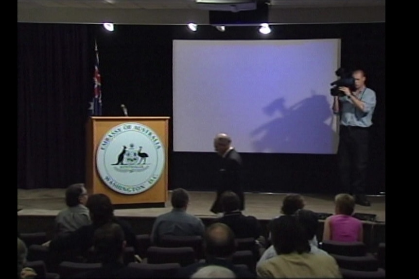 Howard walking to stage in front of media with cameraman on stage filming.