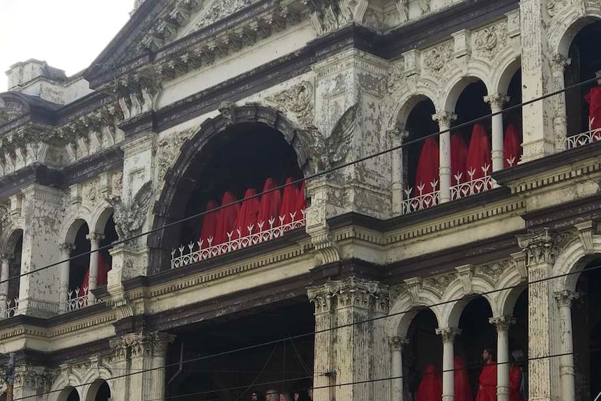 People draped in sheer red fabric lined up along the balcony of a building in Prahran.