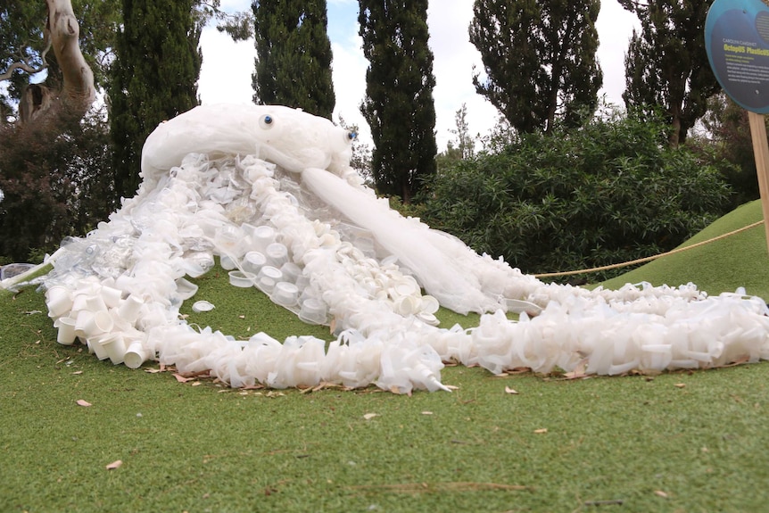 A large white sculpture of a sea creature, made from discarded plastic, sits on a patch of green grass.