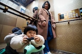 A child leans on a scooter while his mother smiles down at him, inside a lift