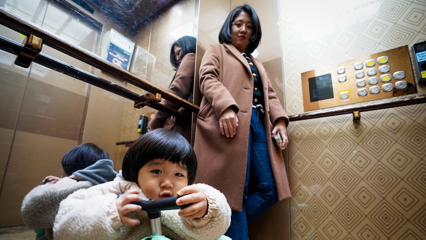 A child leans on a scooter while his mother smiles down at him, inside a lift