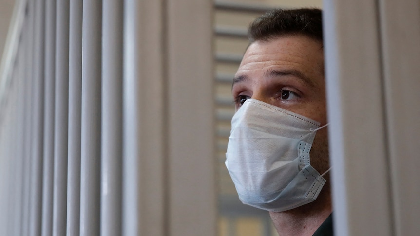 A white man wearing a medical facial mask stands inside a cage during a court hearing.