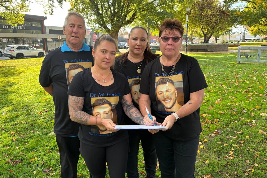 An older man and three women, all wearing matching T-shirts, stand in a park. Two of the women are holding a petition.