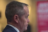 Bill Shorten, captured in side profile, raises his eyebrows while listening. He is wearing a dark suit and blue tie.