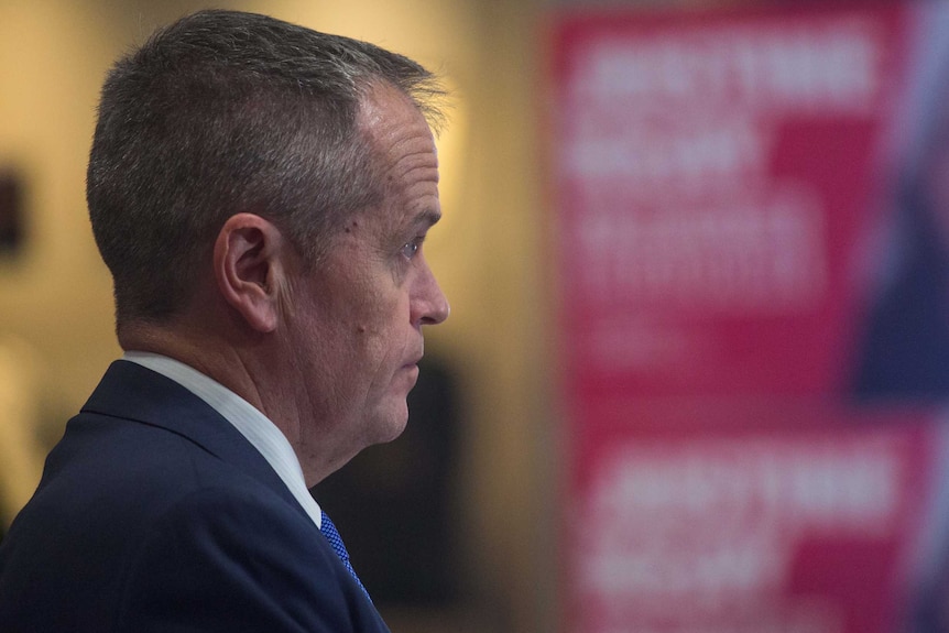 Bill Shorten, captured in side profile, raises his eyebrows while listening. He is wearing a dark suit and blue tie.