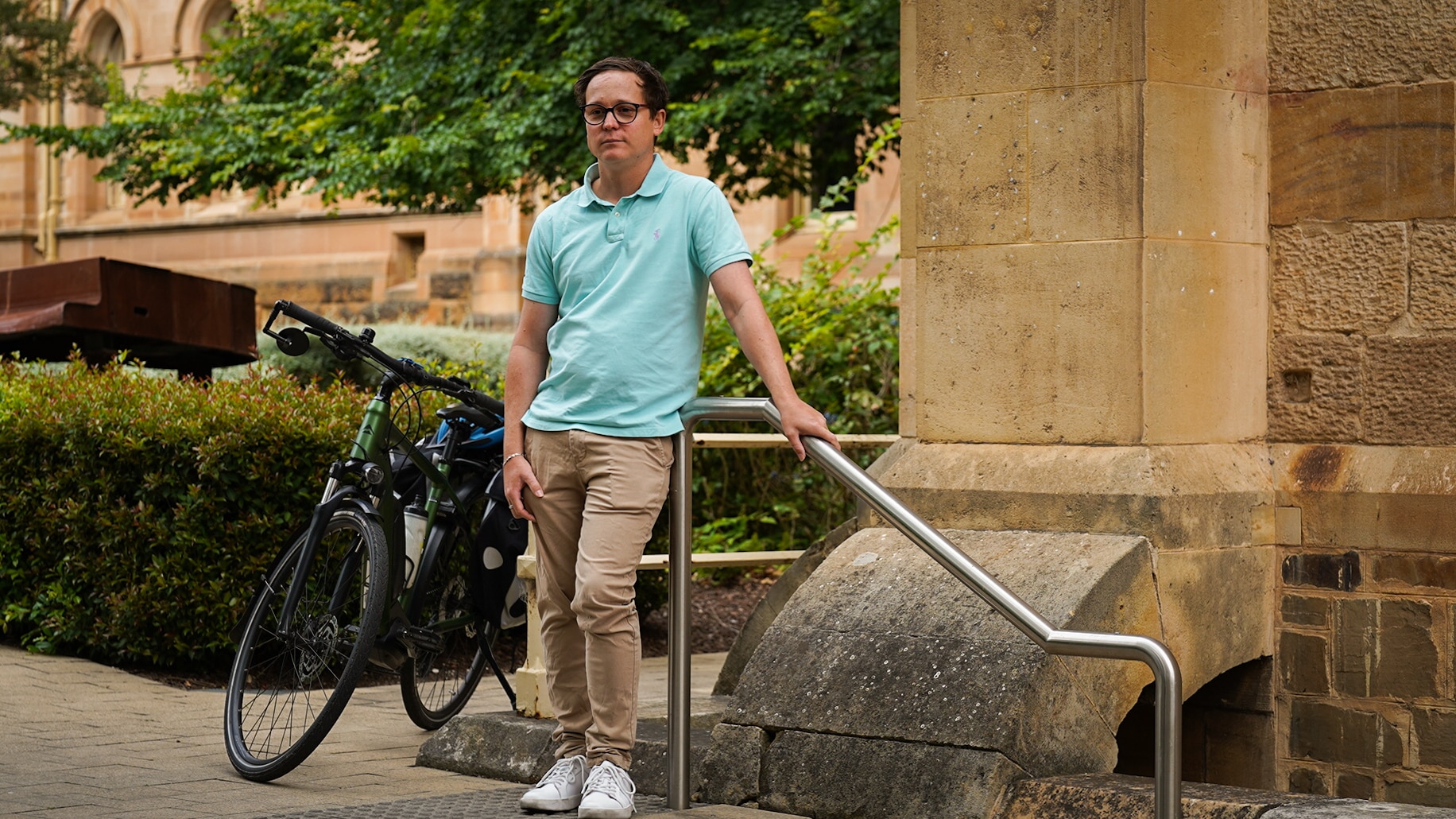 Joshua Bradley veste uma camisa pólo azul clara enquanto apoia a mão em uma grade do lado de fora de um prédio cor de pedra.